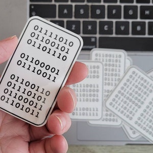May include: A white sticker with black binary code printed on it. The sticker is being held in a hand. There are several other stickers in the background.