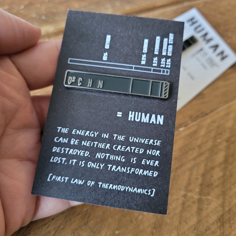 Op de afbeelding: Een zwart-witte emaille pin met een staafdiagram dat de chemische samenstelling van een mens laat zien. De pin heeft de tekst "O2 C H N = HUMAN" en bevat de tekst "DE ENERGIE IN HET UNIVERSUM KAN NIET WORDEN GECRE&Euml;ERD OF VERNIETIGD. NIETS GAAT VERLOREN, HET WORDT ALLEEN GETRANSFORMEERD [EERSTE WET VAN DE THERMODYNAMICA]"