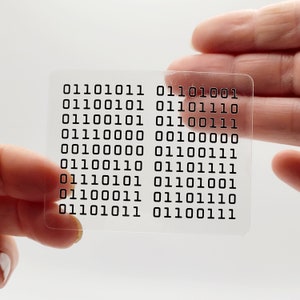 May include: A clear plastic card with a grid of binary code printed on it. The code is made up of 0s and 1s. The card is being held by a hand.