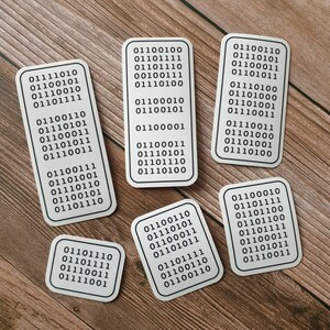 Peut inclure: Six autocollants rectangulaires blancs aux coins arrondis, chacun affichant une s&eacute;quence unique de code binaire noir. Les autocollants sont dispos&eacute;s sur une surface en bois, cr&eacute;ant un contraste entre les &eacute;l&eacute;ments num&eacute;riques et naturels.