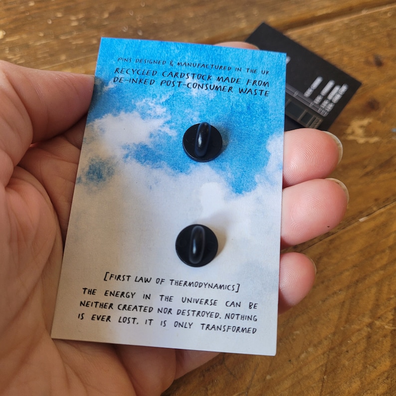 Op de afbeelding: Twee zwarte emaillepins op een blauwe en witte aquarel achtergrond. De pins tonen de tekst "[EERSTE WET VAN DE THERMODYNAMICA] DE ENERGIE IN HET UNIVERSUM KAN NIET WORDEN GECRE&Euml;ERD OF VERNIETIGD. NIETS GAAT OOIT VERLOREN. HET WORDT ALLEEN GETRANSFORMEERD". De achterkant van de kaart luidt "PINS DESIGNED & MANUFACTURED IN THE UK RECYCLED CARDSTOCK MADE FROM DE-INKED POST-CONSUMER WASTE".