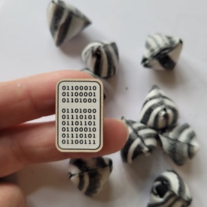 May include: A small wooden card with a series of binary code numbers printed on it. The card is being held by a hand. There are several black and white striped origami fortune tellers scattered around the card.