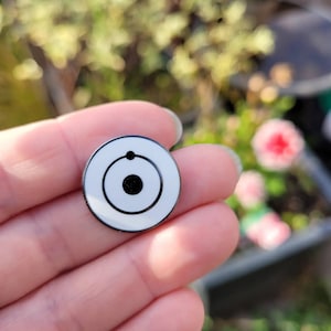 May include: A white enamel pin with a black circle and a smaller black circle inside. The outer circle has a thin black ring around it.