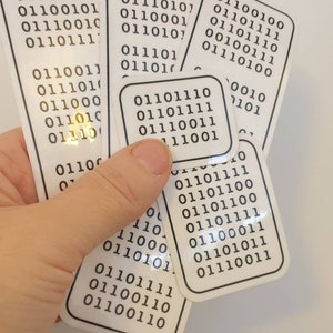 May include: A hand holding several white rectangular stickers with black binary code printed on them. The stickers vary in size and shape, with rounded corners. Some stickers have a gold foil accent.
