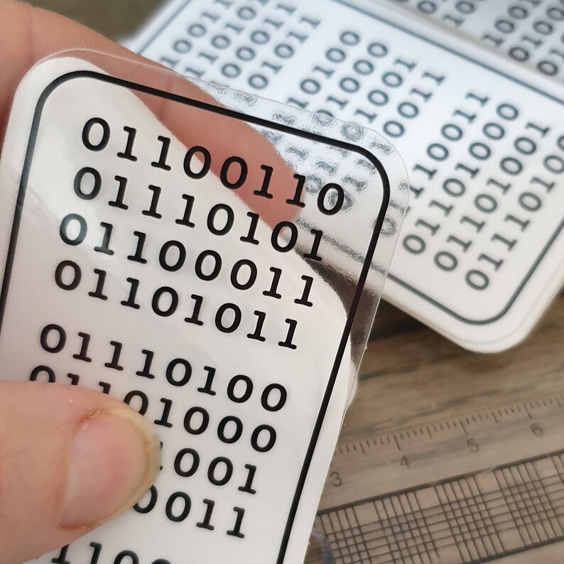 Peut inclure: Autocollants rectangulaires transparents avec du code binaire noir imprim&eacute; sur fond blanc. Les autocollants ont des coins arrondis et un bord noir. Plusieurs autocollants sont visibles, certains nets, d'autres flous.
