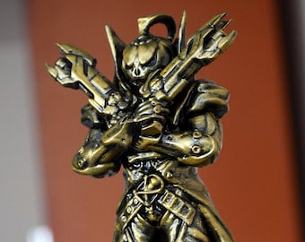 May include: A gold-colored figurine of a skeletal warrior holding two weapons. The warrior is wearing a cape and has a skull-like head. The figurine is standing on a circular base.