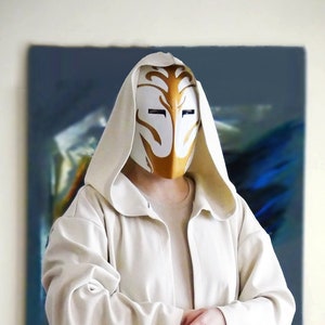 Puede incluir: Una persona lleva una larga túnica blanca con capucha y una máscara blanca y dorada. La túnica tiene mangas largas y un dobladillo ancho y fluido. La persona está de pie en una habitación con una cómoda de madera y un gato en primer plano.