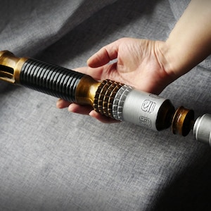 Puede incluir: Un sable de luz desmontado con un mango negro y dorado, una sección central plateada con marcas y una tapa final plateada. El sable de luz está sobre una superficie de tela gris.