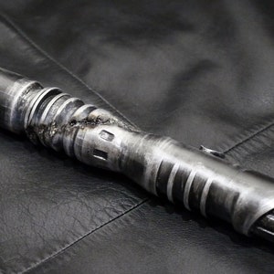 Könnte beinhalten: Ein verwitterter, silberner Lichtschwertgriff mit einer strukturierten Oberfläche und einem schwarzen Griff.