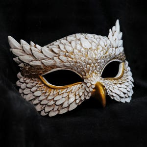May include: A white and gold owl mask with intricate feather detailing. The mask features a gold beak and eye surrounds, with a textured, layered design. The mask is set against a dark background, highlighting its details.