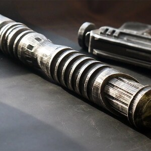 May include: A silver-colored lightsaber prop with a ribbed handle and a blaster pistol prop. The lightsaber has a cylindrical design with detailed grooves and ridges. The blaster pistol is in the background.