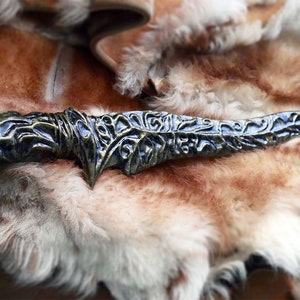 May include: An ornate, antique-style dagger with a bronze and blue finish. The blade features intricate, swirling patterns and a sharp point. The handle is detailed with similar designs. The dagger rests on a backdrop of brown and white fur.