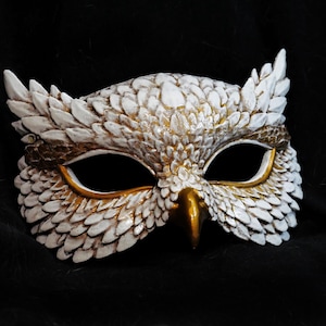 May include: A white and gold owl mask with intricate feather detailing. The mask features large eye openings, a gold beak, and a brown band across the forehead. The mask is designed for costume or decorative purposes.