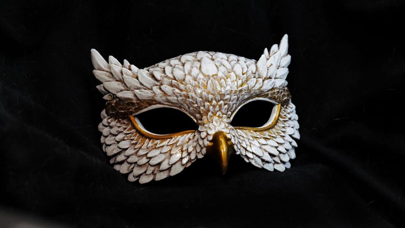 May include: A white and gold owl mask with intricate feather detailing. The mask features large eye openings, a gold beak, and a brown band across the forehead. The mask is designed for costume or decorative purposes.