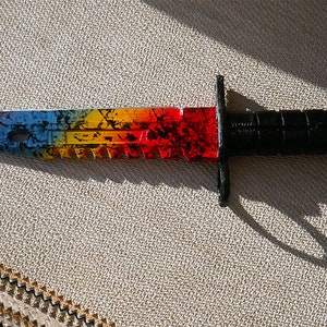 Puede incluir: Un cuchillo de colores arco&iacute;ris con mango negro. La hoja tiene un borde dentado y un patr&oacute;n blanco y negro.