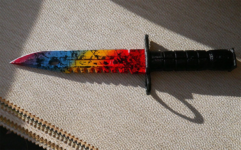 Puede incluir: Un cuchillo de colores arco&iacute;ris con mango negro. La hoja tiene un borde dentado y un patr&oacute;n blanco y negro.