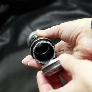 May include: A metal bottle stopper with a cylindrical body and a flat, circular top. The stopper is held in a person's hands, with the top partially detached, revealing the interior.