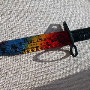 Puede incluir: Un cuchillo de colores arco&iacute;ris con mango negro y hoja dentada. La hoja tiene un aspecto desgastado y envejecido con un esquema de color azul, amarillo, naranja y rojo.