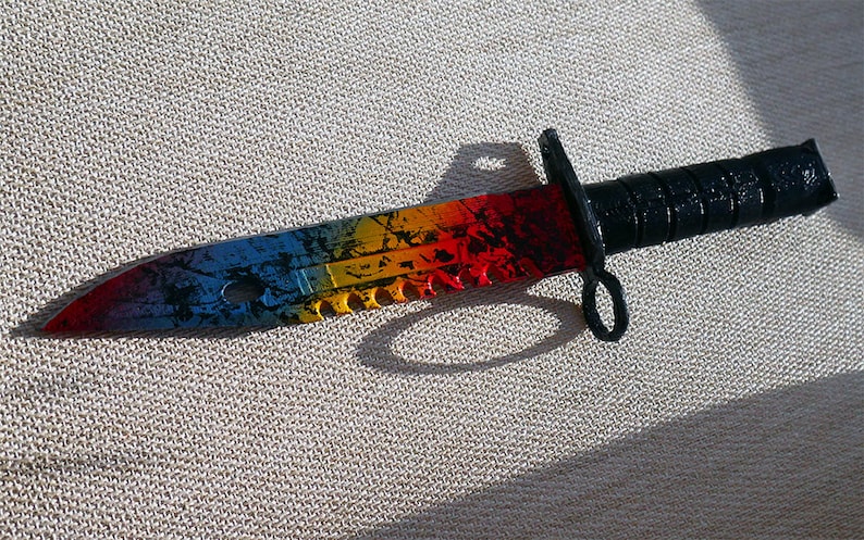 Puede incluir: Un cuchillo de colores arco&iacute;ris con mango negro y hoja dentada. La hoja tiene un aspecto desgastado y envejecido con un esquema de color azul, amarillo, naranja y rojo.