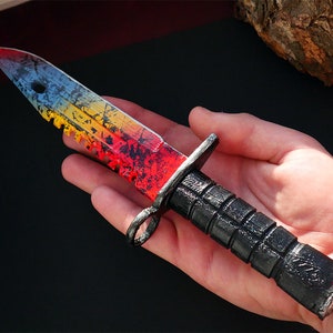Puede incluir: Un cuchillo negro y rojo con una hoja de color arco&iacute;ris y un borde dentado. El cuchillo tiene un mango negro con un agarre texturizado.