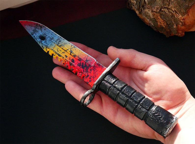 Puede incluir: Un cuchillo negro y rojo con una hoja de color arco&iacute;ris y un borde dentado. El cuchillo tiene un mango negro con un agarre texturizado.