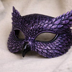 May include: A purple owl mask with a textured, feathered design. The mask has black eye openings and a black beak. The mask is made of a hard material and is designed to be worn on the face.