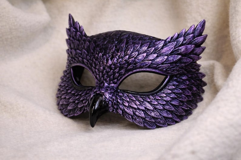 May include: A purple owl mask with a textured, feathered design. The mask has black eye openings and a black beak. The mask is made of a hard material and is designed to be worn on the face.