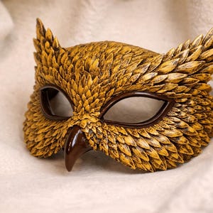 May include: A handcrafted owl mask with a textured, golden-yellow finish. The mask features dark brown eye openings and a beak, with detailed feather-like patterns. The mask is designed to be worn and is set against a soft, white background.
