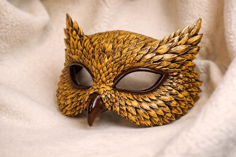 May include: A handcrafted owl mask with a textured, golden-yellow finish. The mask features dark brown eye openings and a beak, with detailed feather-like patterns. The mask is designed to be worn and is set against a soft, white background.