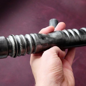 May include: A metallic gray lightsaber prop with a detailed, textured design. The prop features a curved blade opening and a ribbed handle. The lightsaber is held against a dark purple background.