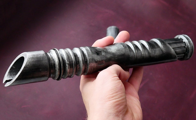 May include: A metallic gray lightsaber prop with a detailed, textured design. The prop features a curved blade opening and a ribbed handle. The lightsaber is held against a dark purple background.