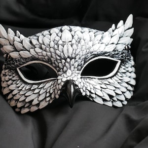 May include: A white and black owl mask with a detailed feather design. The mask has large eye openings and a beak, perfect for costume parties or theatrical performances. The mask is made of a durable material.