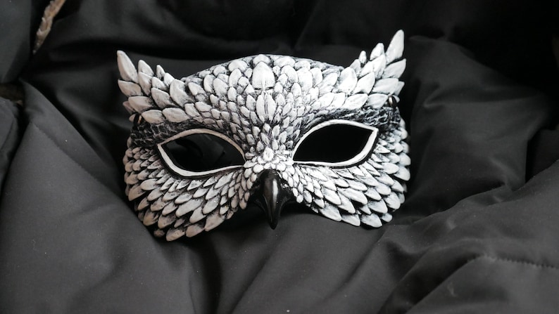 May include: A white and black owl mask with a detailed feather design. The mask has large eye openings and a beak, perfect for costume parties or theatrical performances. The mask is made of a durable material.