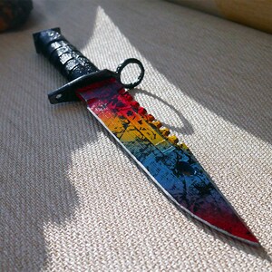 Puede incluir: Un cuchillo de arco&iacute;ris con mango negro y hoja dentada. El cuchillo tiene un patr&oacute;n de arco&iacute;ris en la hoja, con colores rojo, naranja, amarillo, verde, azul, &iacute;ndigo y violeta.