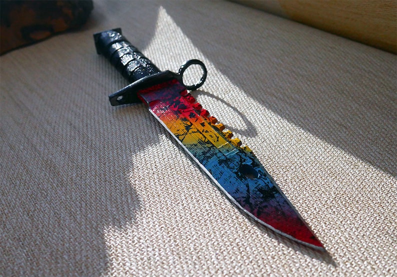 Puede incluir: Un cuchillo de arco&iacute;ris con mango negro y hoja dentada. El cuchillo tiene un patr&oacute;n de arco&iacute;ris en la hoja, con colores rojo, naranja, amarillo, verde, azul, &iacute;ndigo y violeta.