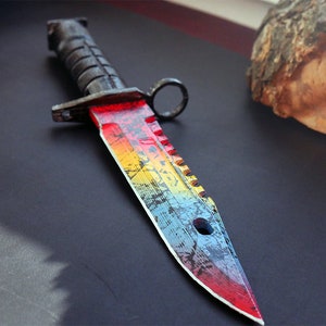 Puede incluir: Un cuchillo de color arco&iacute;ris con mango negro y hoja dentada. La hoja tiene un aspecto desgastado con una pintura de arco&iacute;ris.