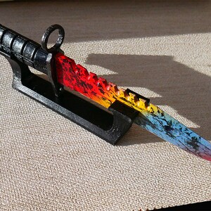 Puede incluir: Un cuchillo de colores arco&iacute;ris con mango negro y hoja dentada se muestra en un soporte negro. El cuchillo tiene un degradado de color rojo, naranja, amarillo, azul y morado.