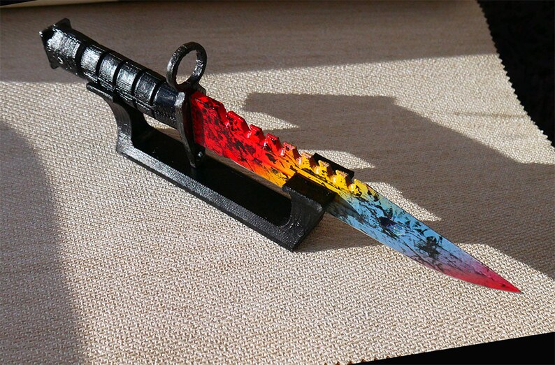 Puede incluir: Un cuchillo de colores arco&iacute;ris con mango negro y hoja dentada se muestra en un soporte negro. El cuchillo tiene un degradado de color rojo, naranja, amarillo, azul y morado.