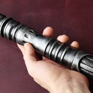 May include: A metallic gray lightsaber prop with a textured finish. The prop features a cylindrical design with ridged sections and a detailed emitter. The lightsaber is held in a hand, showcasing its size and design.