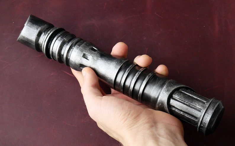 May include: A metallic gray lightsaber prop with a textured finish. The prop features a cylindrical design with ridged sections and a detailed emitter. The lightsaber is held in a hand, showcasing its size and design.