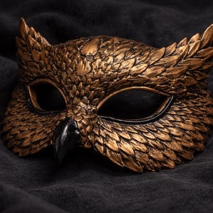 May include: A detailed, gold-colored owl mask with a black beak and eye surrounds. The mask has a textured, feathered appearance and is set against a dark, textured background. This decorative mask is suitable for costume parties or theatrical events.