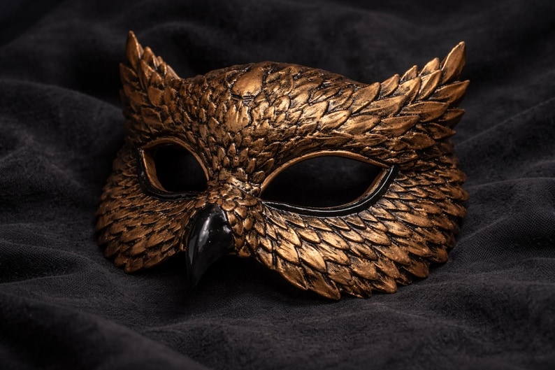 May include: A detailed, gold-colored owl mask with a black beak and eye surrounds. The mask has a textured, feathered appearance and is set against a dark, textured background. This decorative mask is suitable for costume parties or theatrical events.