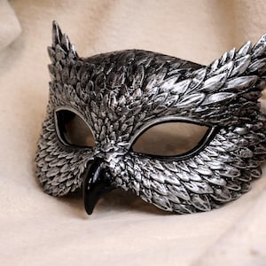 May include: A silver and black owl mask with detailed feather textures. The mask features large eye openings and a prominent beak. The mask is designed to be worn and is likely for costume or decorative purposes.