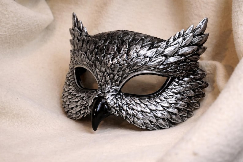 May include: A silver and black owl mask with detailed feather textures. The mask features large eye openings and a prominent beak. The mask is designed to be worn and is likely for costume or decorative purposes.