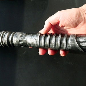 May include: A silver and black lightsaber prop with a detailed, textured design. The hilt features ridged sections and a curved emitter. The prop is held in a hand against a dark background, showcasing its metallic appearance.
