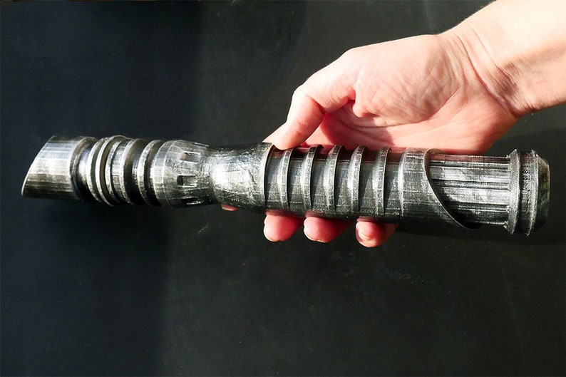 May include: A silver and black lightsaber prop with a detailed, textured design. The hilt features ridged sections and a curved emitter. The prop is held in a hand against a dark background, showcasing its metallic appearance.