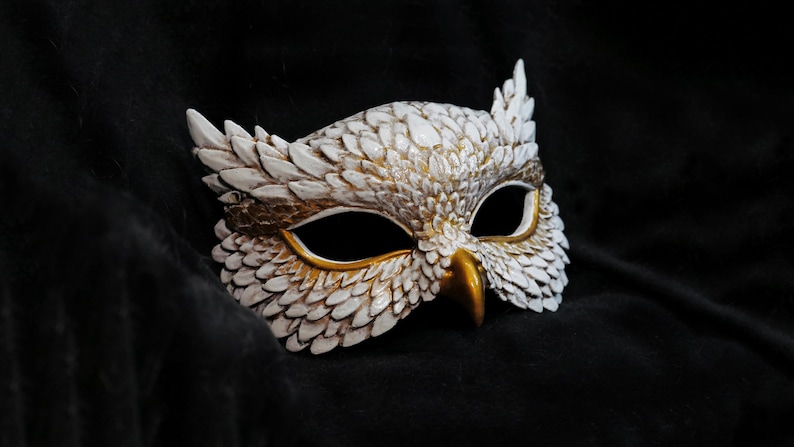 May include: A white and gold owl mask with intricate feather detailing. The mask features a gold beak and eye surrounds, with a textured, layered design. The mask is set against a dark background, highlighting its details.