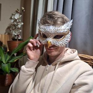 May include: An owl mask with white and gold details. The mask features a detailed feather design, a gold beak, and is worn by a person wearing a beige hoodie. The mask is likely for a costume or decorative purpose.