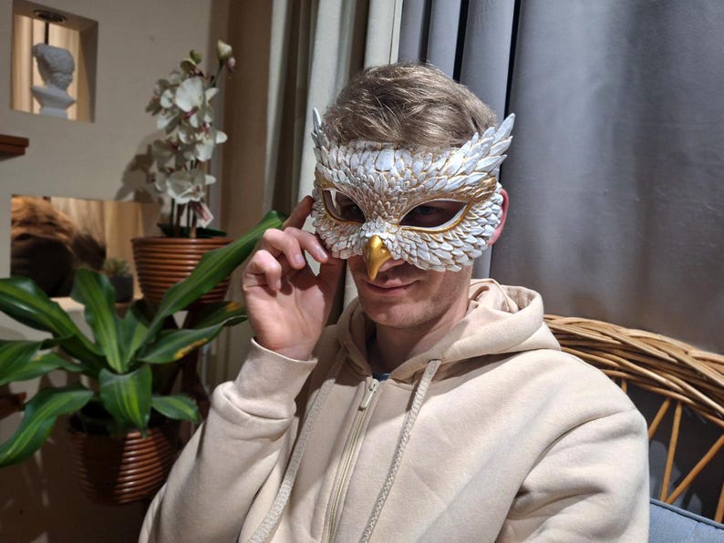 May include: An owl mask with white and gold details. The mask features a detailed feather design, a gold beak, and is worn by a person wearing a beige hoodie. The mask is likely for a costume or decorative purpose.
