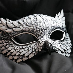 May include: A white and black owl mask with detailed feather textures. The mask has large eye openings and a prominent beak. The mask is resting on a dark, textured fabric, creating a dramatic contrast.